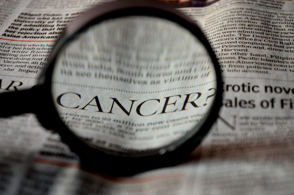 Close-up of a magnifying glass over a newspaper, highlighting the word 'CANCER?'.