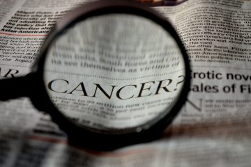 Close-up of a magnifying glass over a newspaper, highlighting the word 'CANCER?'.