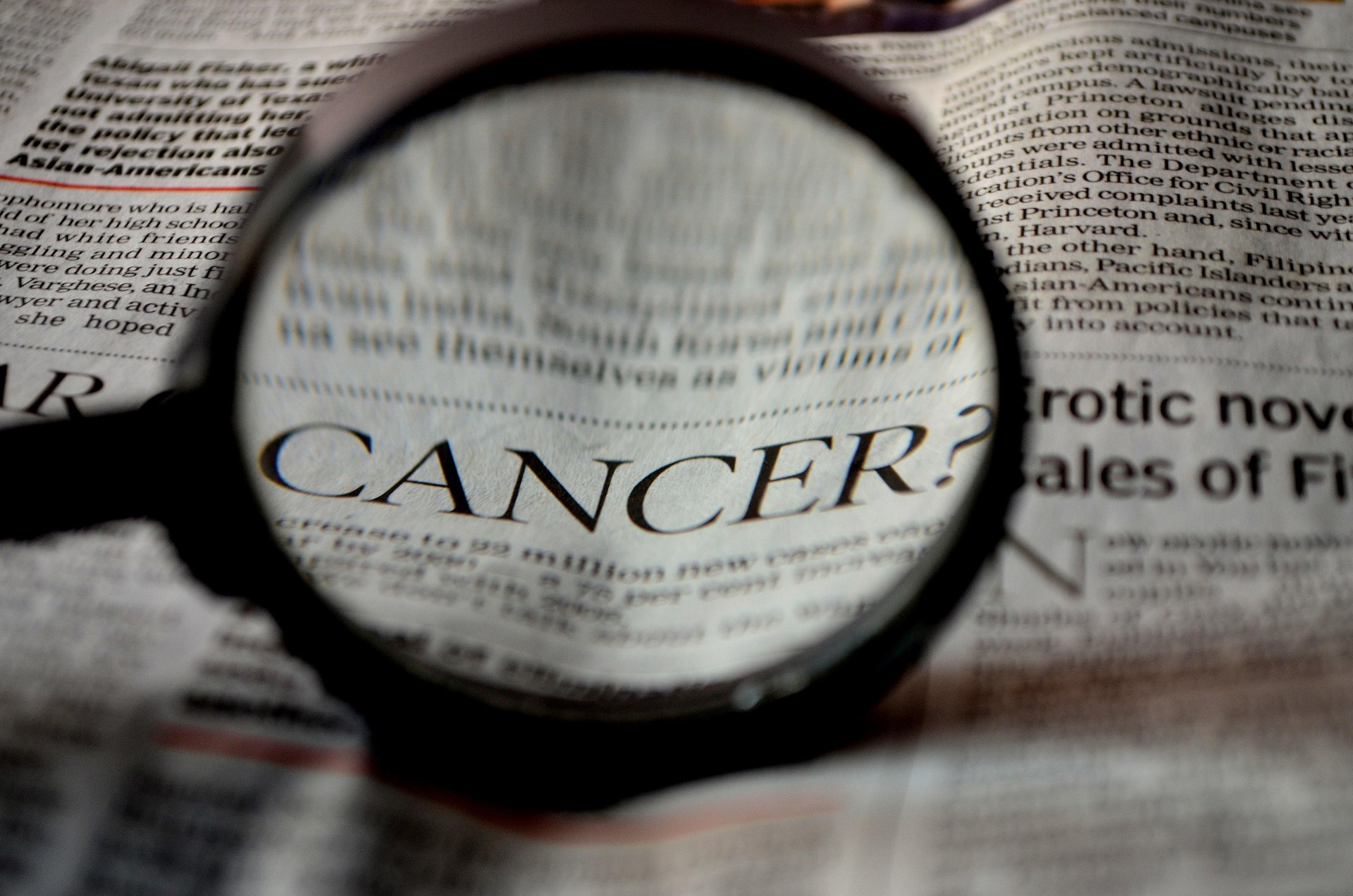 Close-up of a magnifying glass over a newspaper, highlighting the word 'CANCER?'.