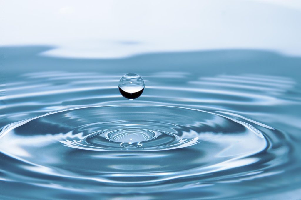 A single water droplet hovering just above blue ripples on a calm water surface.