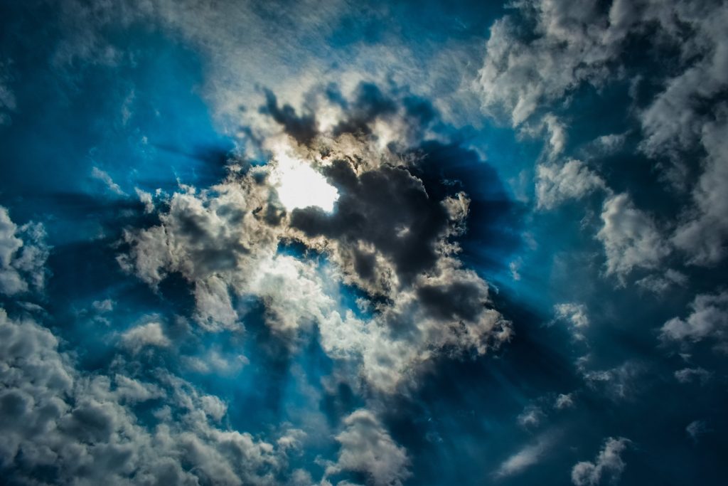 Sun breaking through dark clouds, with bright rays streaming across a dramatic blue sky.