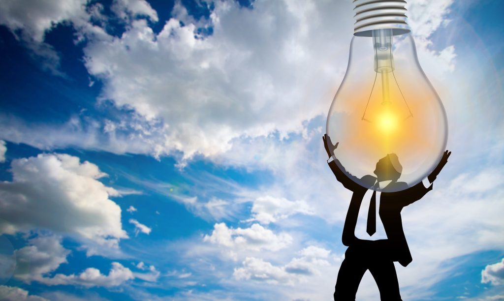 Silhouetted figures raise a large glowing light bulb against a blue, cloudy sky, symbolizing a bright idea or innovation.