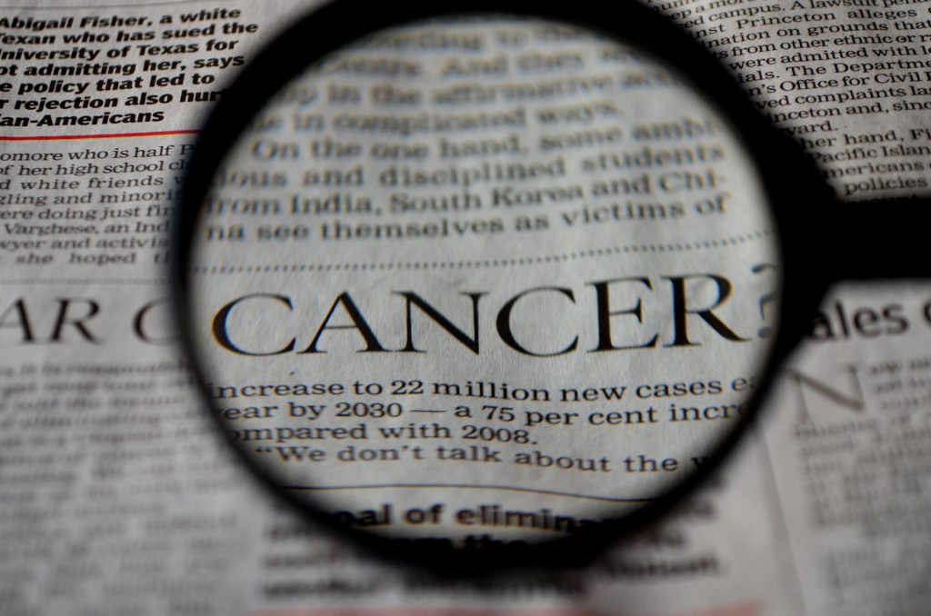 Magnifying glass over a newspaper, focusing on the word 'CANCER' in bold print.