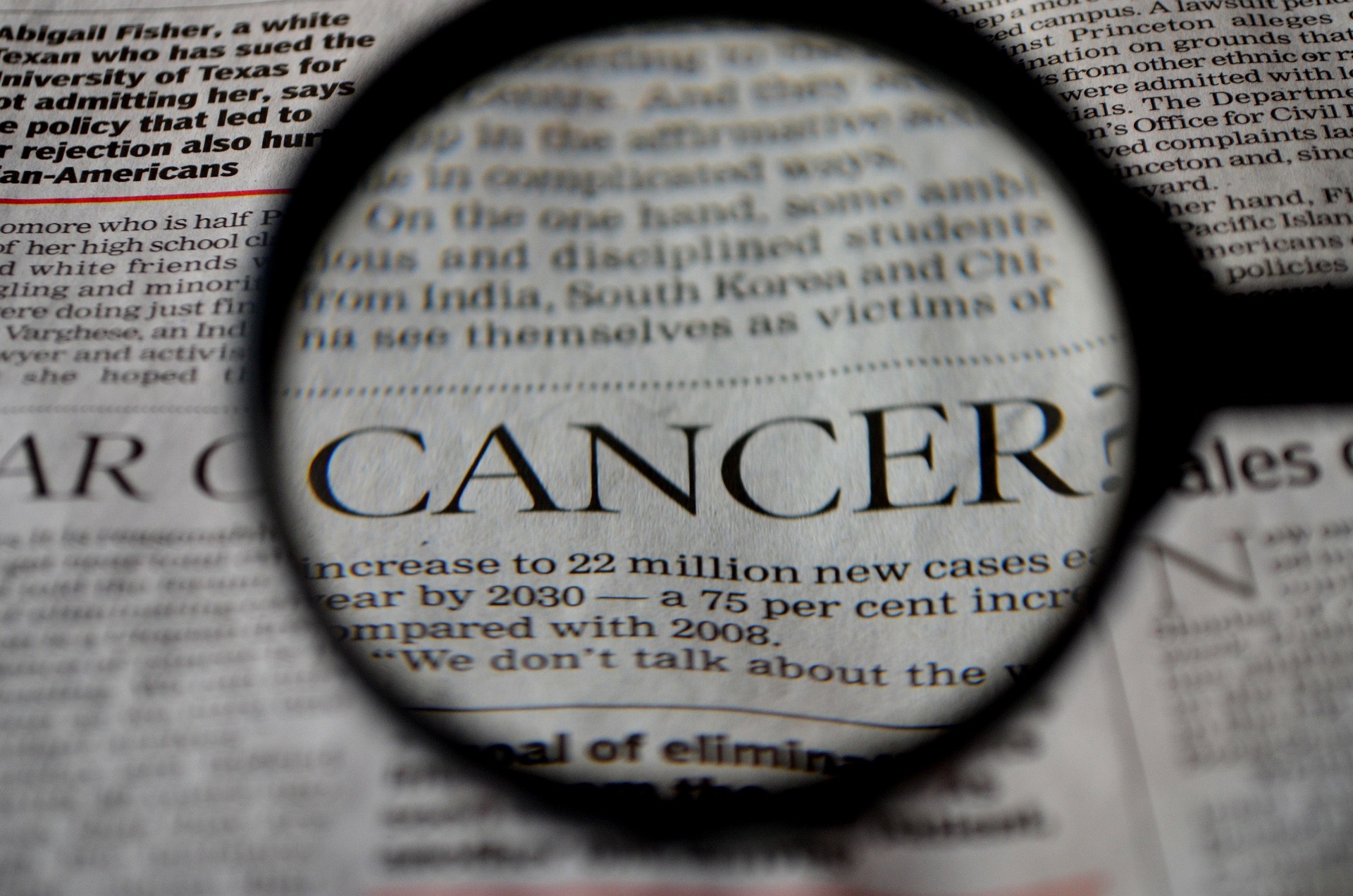 Magnifying glass over a newspaper, focusing on the word 'CANCER' in bold print.