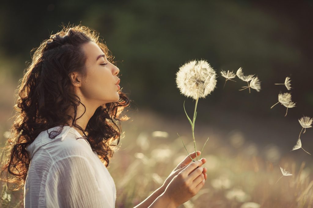 Woman with curly hair blowing a dandelion seed head in a sunlit field at golden hour.