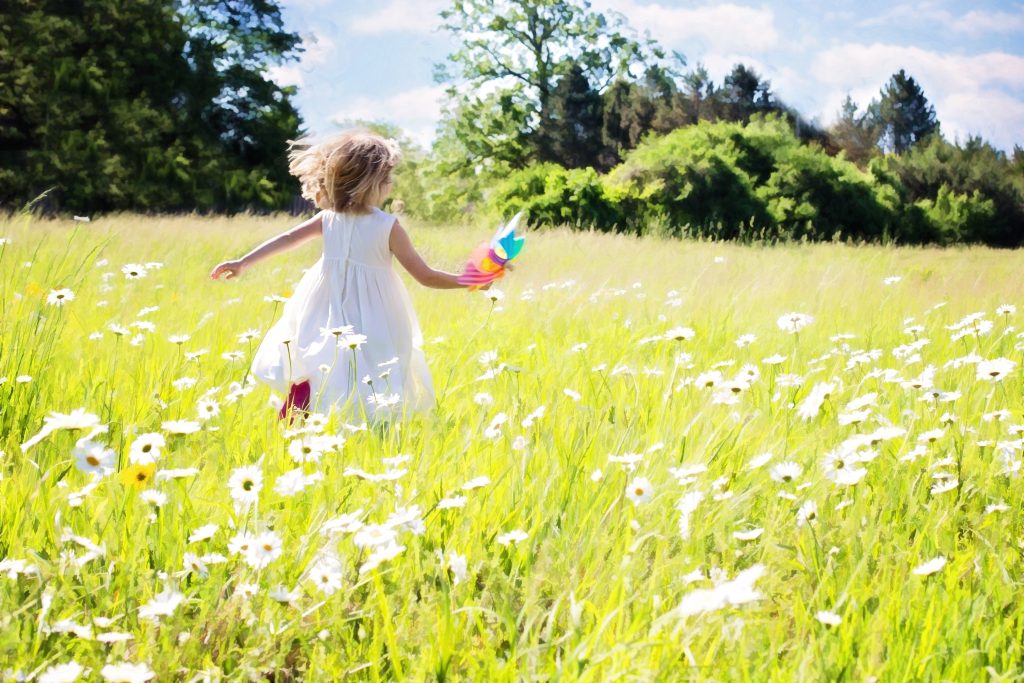 Young girl in a white dress runs through a sunny meadow full of white daisies, holding a colorful pinwheel in her hand.