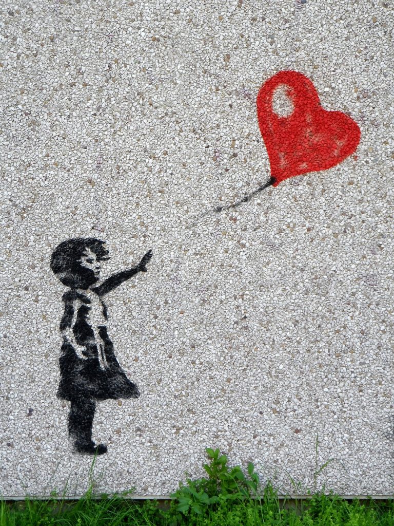Black silhouette of a person aiming a gun toward a red heart graffiti on a speckled wall, with grass at the base.